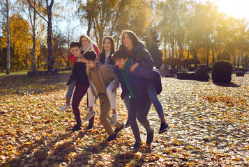 Bunch of funny friends having fun while walking all together in a sunny autumn park. Group of several happy funny cheerful young people piggy backing their girlfriends, running and having a race
