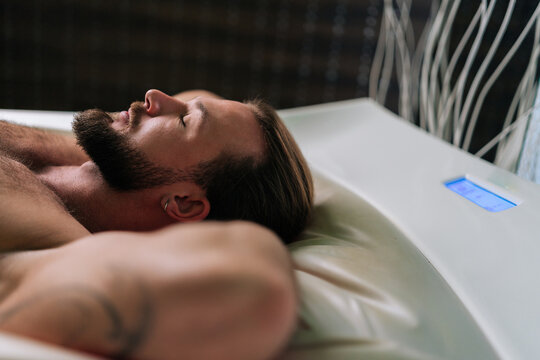 Closeup side view face of relaxed young man with earring in ear lying relaxing on electric massage bed with closed eyes, enjoying spa treatment at spa salon. Muscular male having rest enjoying weekend
