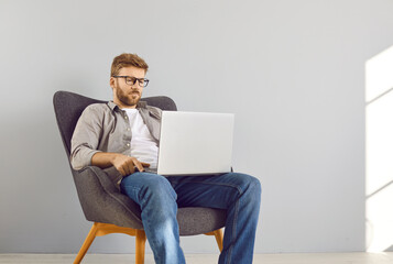 Portrait of a young bearded businessman sitting on chair in empty space with laptop working or...
