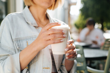 Close-up of woman in casual clothes and light manicure holding white disposable cup of coffee while...