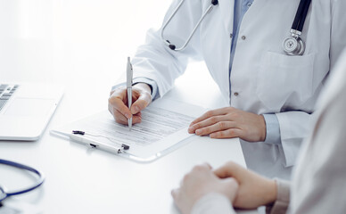 Doctor and patient sitting at the table in clinic. The focus is on female physician's hands filling up the medication history record form or checklist, close up. Medicine concept