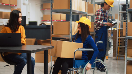 Fototapeta na wymiar Depot employee with disability taking boxes off of racks in warehouse space, organizing products on shelves. Young adult in wheelchair talking to owner about business development and shipment.