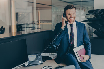 Businessman in formal wear, speaking on his office desk phone while sitting on top of table