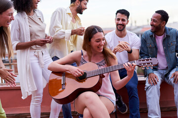 Group of people reunited in a terrace. Caucasian woman playing a guitar surrounded by her friends enjoying in a terrace, singing and laughing outdoors.