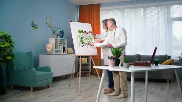 an elderly man with his old wife learn new skill in retirement and paint picture with paints and brushes on canvas using an easel in room