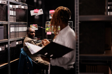Private detective helping partner searching crime case information, standing near shelf with folder. African american investigators analyzing evidence and report in agency office