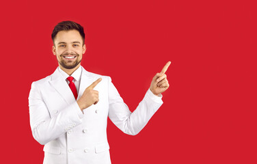 Portrait of smiling young Caucasian man in suit isolated on red studio background point at blank copy space aside. Happy handsome male in costume recommend sale deal or promotion offer.