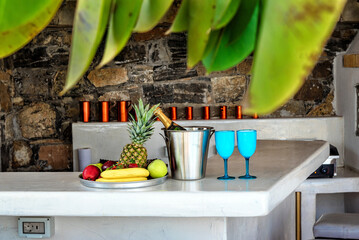 Relax and holiday concept, white bar counter with an ice basket containing a bottle, two blue glasses, and a fruit plate with bananas, apples, and pineapple. 