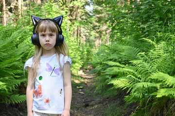 A little girl with long blond hair stands in the woods and listens to the uzyka in headphones in...