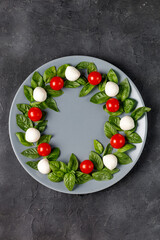 Fototapeta na wymiar Caprese salad in the form of a Christmas wreath. Festive tomato mozzarella and basil appetizer on grey plate.