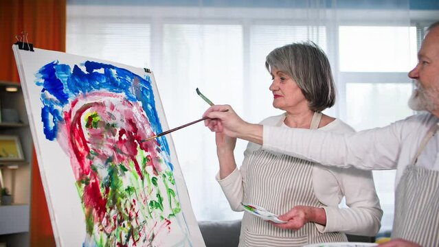 talented pensioners, an elderly man and his wife develop new drawing skills and paint a picture together with paints and brushes using an easel at home