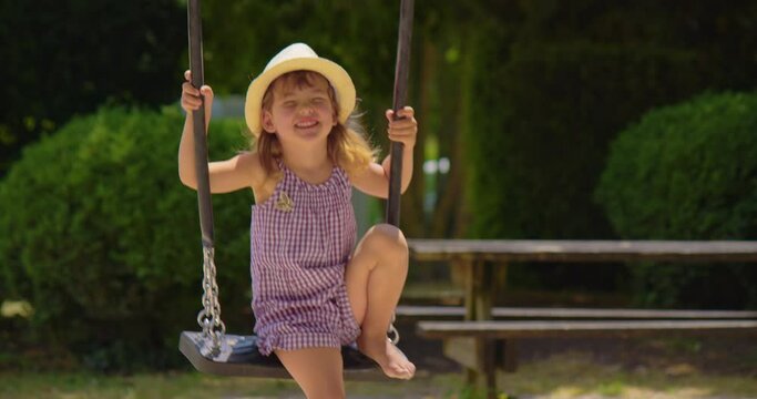 A little girl of three years in a hat and dress rides on a swing in the park in summer
