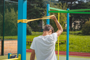Fototapeta premium Goal-oriented teenager working on his body using resistance rubber performs strength training. Preparing for the sports season. Exercising with your own body. Outdoor workout playground. Biceps