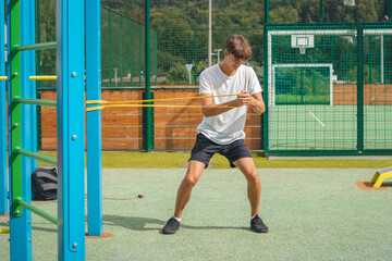 Focused teenager working on his body using resistance rubber performs strength training. Preparing for the season. Exercising with your own body. Outdoor workout playground. Oblique abdominal muscles