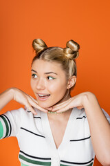 Cheerful and blonde teenager with colorful makeup and hairstyle holding hands near chin and looking...