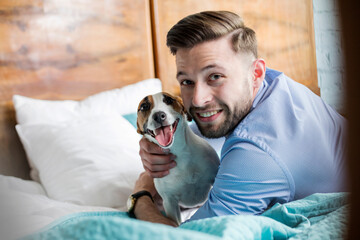 Portrait smiling man petting Jack Russell Terrier dog on bed - obrazy, fototapety, plakaty