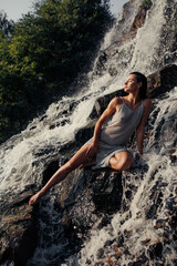 Young wet woman sitting on rock near waterfall between water flows.