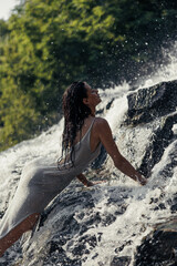 Young woman in wet dress lying on rock in waterfall.