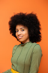 Obraz na płótnie Canvas Portrait of fashionable teen african american girl with bold makeup wearing green dress and smiling at camera isolated on orange, youth culture and generation z concept