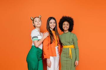 Group of interracial teenage models in stylish outfit posing and smiling at camera while standing together on orange background, trendy generation z concept, friendship and companionship