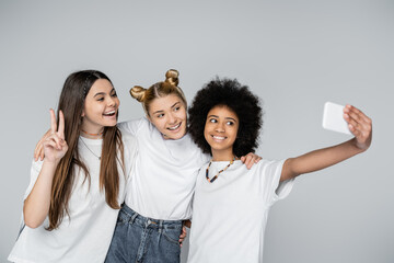 Smiling teen african american girl in white t-shirt taking selfie on smartphone with girlfriends hugging each other isolated on grey, teenagers bonding over common interest, friendship