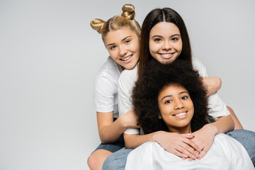 Joyful interracial teenage girlfriends in white t-shirts looking at camera while hugging each other and posing isolated on grey, energetic teenage friends spending time, copy space