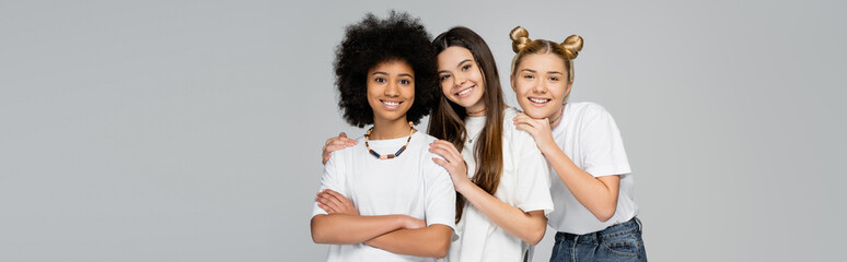 Positive multiethnic teen girlfriends in casual t-shirts and jeans hugging each other and looking at camera together isolated on grey, lively teenage girls concept, banner with copy space