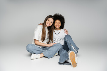 Smiling brunette teen girl in white t-shirt and jeans hugging african american girlfriend while sitting together and looking at camera on grey background, lively teenage girls concept