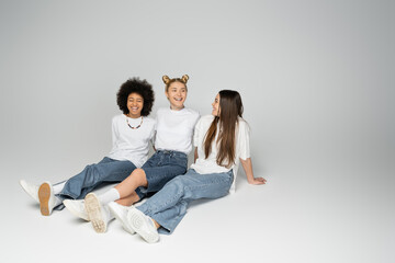 Full length of positive and multiethnic teenage girls in white t-shirts and blue jeans talking while sitting and spending time together on grey background, multiethnic teen models concept