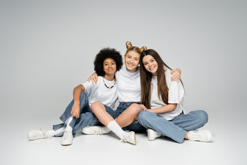 Joyful and multiethnic teenage girls in stylish white t-shirts and blue jeans hugging and looking at camera while sitting together on grey background, multiethnic teen models concept