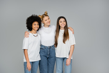 Fototapeta premium Smiling blonde teenage girl in white t-shirt and blue jeans hugging multiethnic friends and looking away while standing isolated on grey, teenage friends having fun together