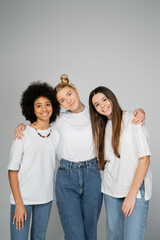 Fototapeta premium Stylish and cheerful interracial teenage girls in white t-shirts and blue jeans hugging and looking at camera while standing isolated on grey, teenage friends having fun together