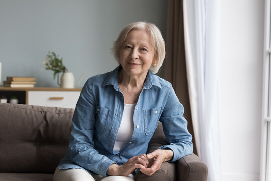 Calm retired mature woman in casual clothes resting alone seated on comfortable sofa in living room smile looking at camera at own modern apartment. Baby-boomer generation female portrait, retirement