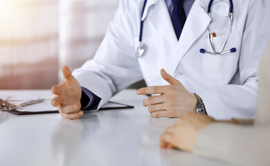 Unknown male doctor and patient woman discussing something while sittingin a darkened clinic, glare of light on the background. Close-up of hands