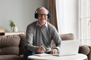 Positive senior man in headphones looking at camera studying online at home with laptop, get...