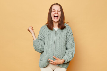 Positive smiling young pregnant woman wearing knitted sweater standing isolated over beige background rejoicing celebrating success clenched her fist.