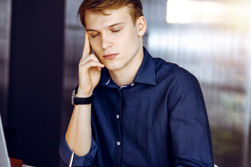 Young blond businessman feeling stress at workplace in a darkened office, glare of light on the background. Startup business means working hard and out of time