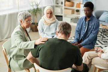 Rear view of mature stressed man with post traumatic syndrome sitting among intercultural people and psychologist comforting him