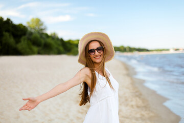 Fototapeta na wymiar Happy smiling woman in free happiness bliss on ocean beach standing with a hat, sunglasses, and open hands. Portrait of a multicultural female model in white summer dress enjoying nature during travel