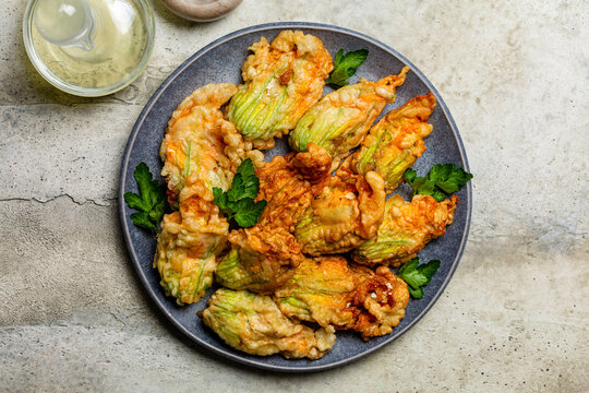 Plate With Fried In A Batter Zucchini Flowers Stuffed With Ricotta Cheese And Parsley. Raw And Roasted Courgette Or Pumpkin Flowers. Italian Dish Fiori Di Zucca In Pastella. Stone Background.