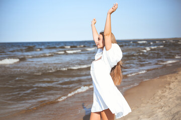 Happy smiling beautiful woman on the ocean beach standing in a white summer dress, raising hands