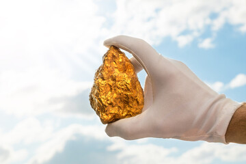 Golden nugget in a man's hand in a white glove raised to the sky. The concept of wealth and success in the financial sector. Treasures, investments. Mining industry.