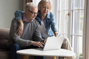 Mature couple search last minute tours, buy tickets, choose electronic services and goods, making order through e-services on laptop. Older generation using modern tech, easy, comfort purchases online