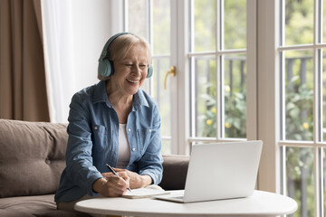Carefree smiling female pensioner listen video lesson on computer use headphones, acquires new abilities, skill, gain knowledge engaged in self-education, enjoy opportunity new earning in retirement