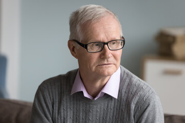 Head shot portrait of concerned gloomy serious mature man in glasses sit on sofa alone at home,...