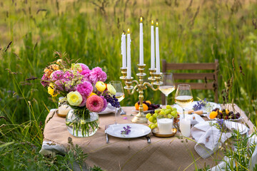 Beautiful countryside decor for romantic dinner or small wedding ceremony in a field. Table decorated with ranunculi bright flowers, cheese, grapes, chandelier with burning candles. Sunset golden hour