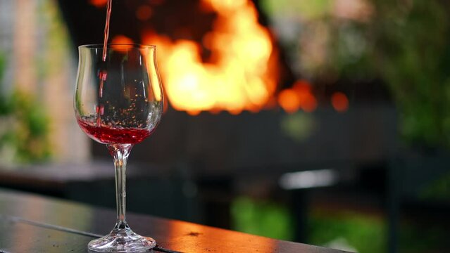 Close-up glass on the left at background of bbq fire and red wine pouring in slow motion. Closeup delicious alcohol beverage on barbecue picnic at night outdoors