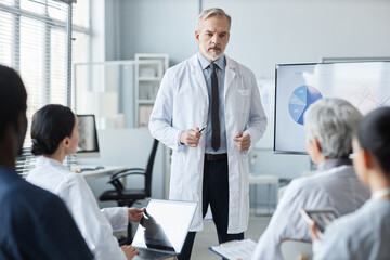 Confident mature male general practitioner in lab coat making speech or presentation while standing...