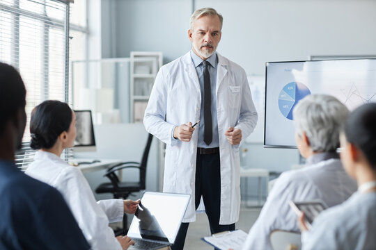 Confident mature male general practitioner in lab coat making speech or presentation while standing by whiteboard in front of audience