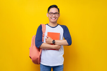 Smiling attractive young Asian man student wearing casual clothes bag standing confident while holding book isolated on yellow background. Education in high school university college concept